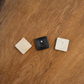 Three Shelly devices—two beige and one black (Shelly BLU H&T ZB Black)—are arranged side by side on a wooden surface, each featuring a circular button with a smaller dot above it.