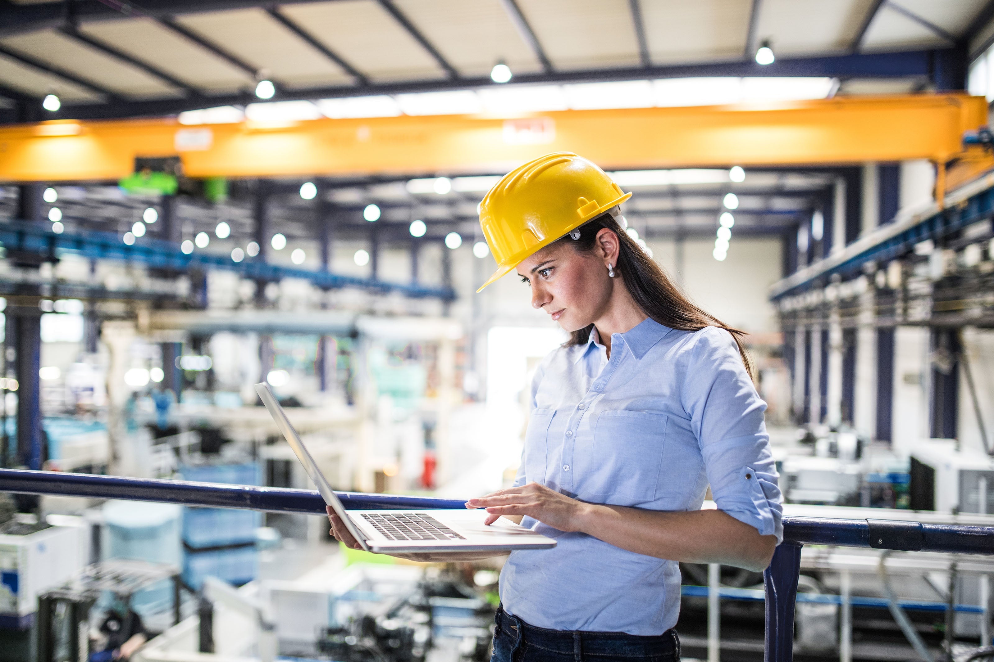 A woman wearing a yellow hard hat and blue shirt stands in a factory, using a laptop. Industrial equipment and machinery are visible in the background.