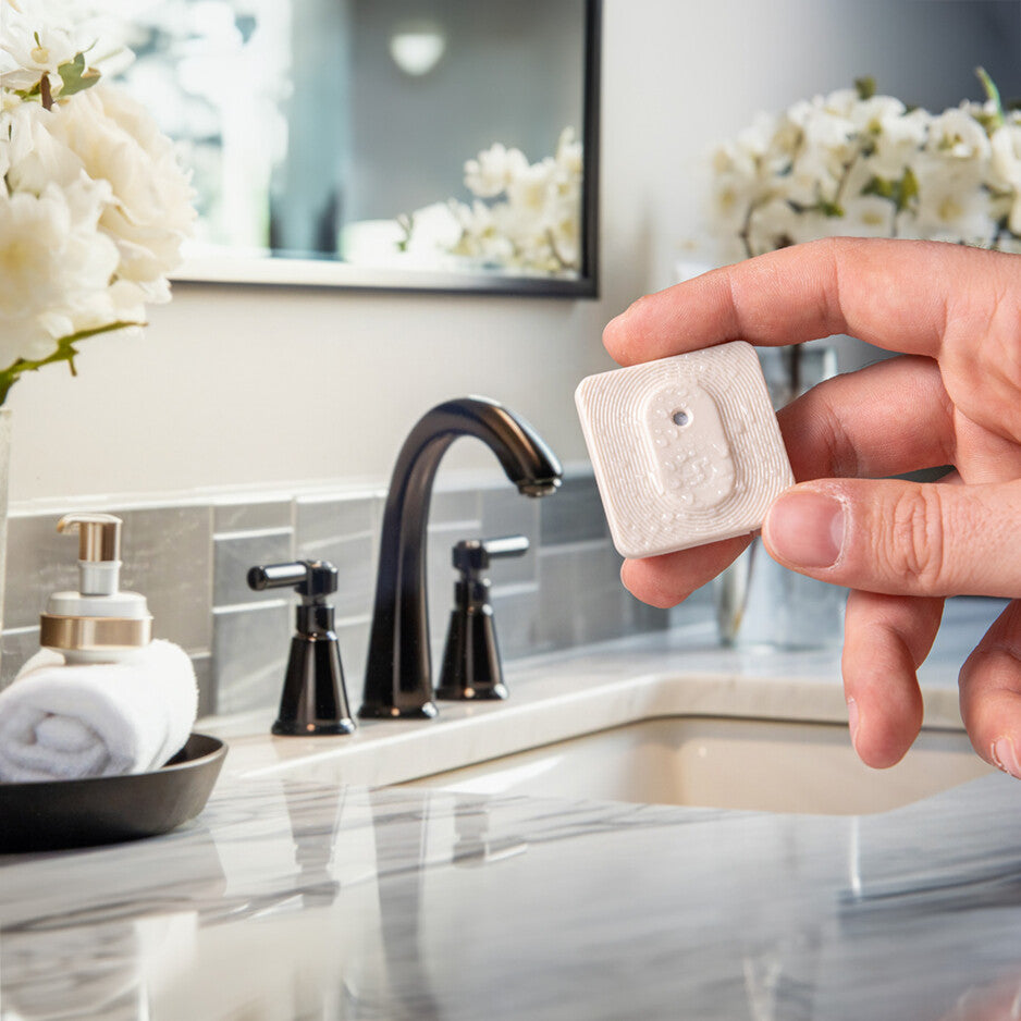 A hand holds the Shelly BLU H&T ZB Ivory by Shelly Europe above a marble bathroom countertop with a black faucet, towel, soap dispenser, and white flowers in the background.