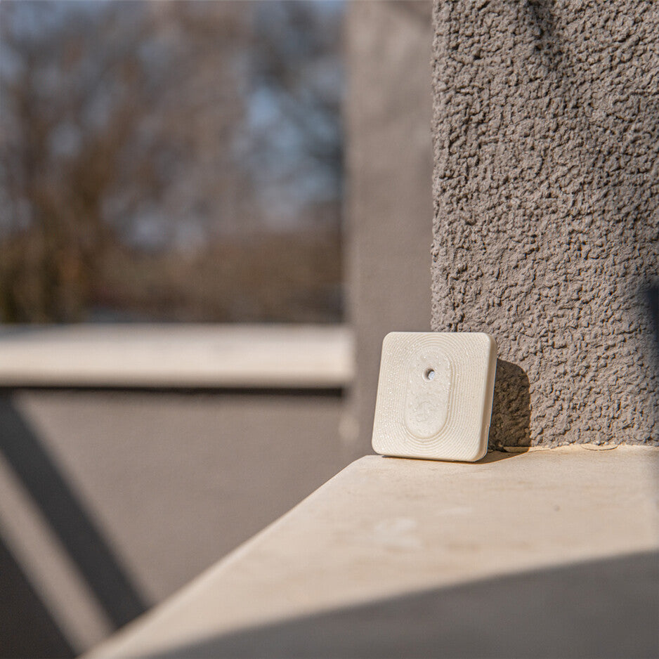 The Shelly Europe Shelly BLU H&T ZB Ivory, a compact square off-white sensor, is mounted on a textured gray outdoor wall with sunlight casting shadows and blurred trees in the background.