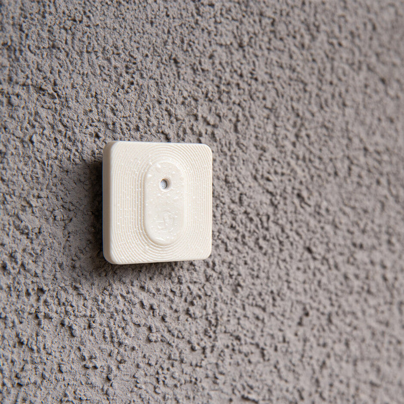 The Shelly BLU H&T ZB Ivory by Shelly Europe is a compact, white, square plastic device with rounded edges and a central oval feature featuring a small hole, mounted on a textured gray wall.
