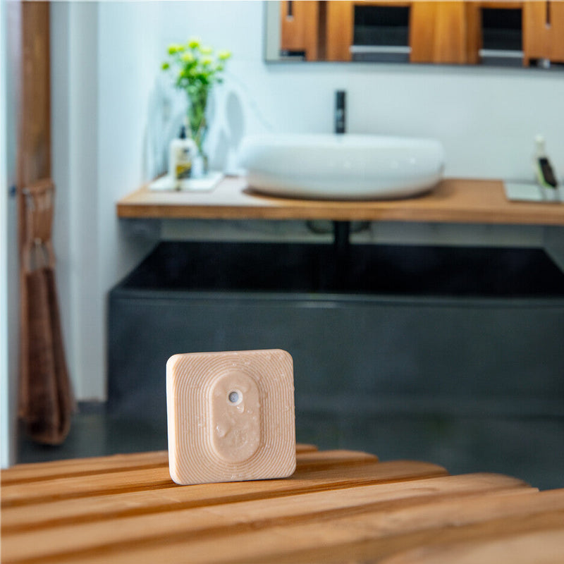 A Shelly Shelly BLU H&T ZB Mocha motion sensor in beige sits on a wooden bathroom counter, with a white sink and vase of yellow flowers visible in the blurred background.