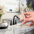 A hand holds the Shelly BLU H&T ZB Mocha by Shelly near a bathroom sink with a marble countertop, black faucet, soap dispenser, rolled towel, and white floral decorations in the background.