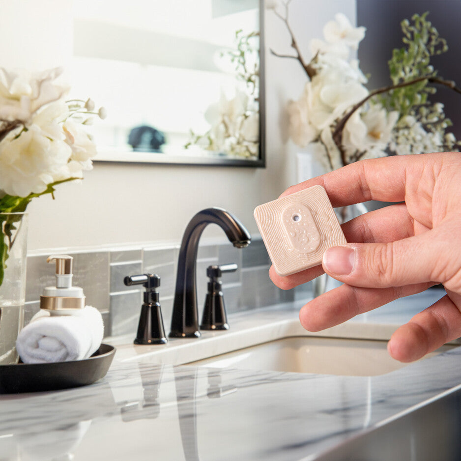 A hand holds the Shelly BLU H&T ZB Mocha by Shelly near a bathroom sink with a marble countertop, black faucet, soap dispenser, rolled towel, and white floral decorations in the background.