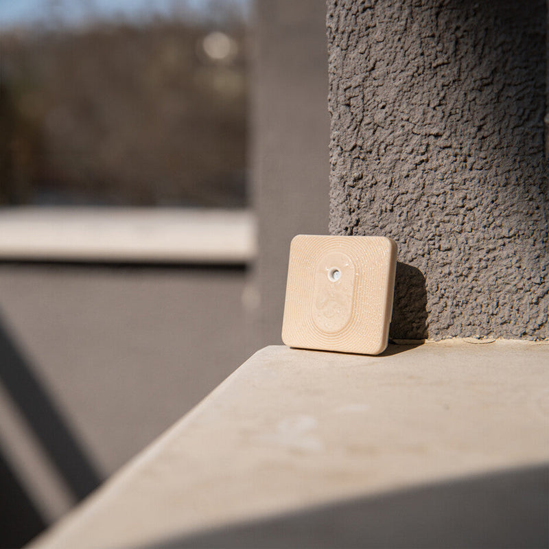 The Shelly Shelly BLU H&T ZB Mocha, a small square light-colored device with a circular button, is mounted on a textured gray wall of a sunny balcony with a blurred outdoor background.