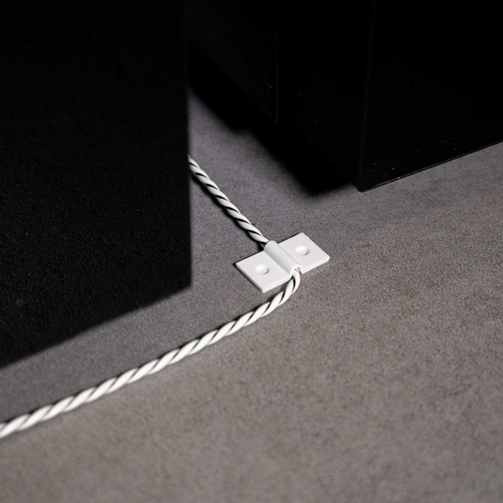 A white cable secured to a grey floor using two Shelly Europe Shelly Cable Bracket clips, running alongside black furniture.
