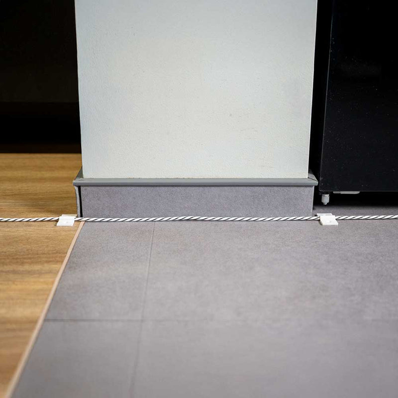 A white cable, secured by Shelly Europe’s Shelly Cable Bracket 10-pack, runs along the floor between wooden and gray tile sections beside a light wall and a black appliance.