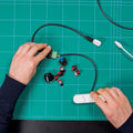 A person assembles electronic components for an electronics project, including the Shelly USA Micro USB to 5 Terminal Add (Shelly Pill), while holding cables and parts over a green gridded cutting mat.