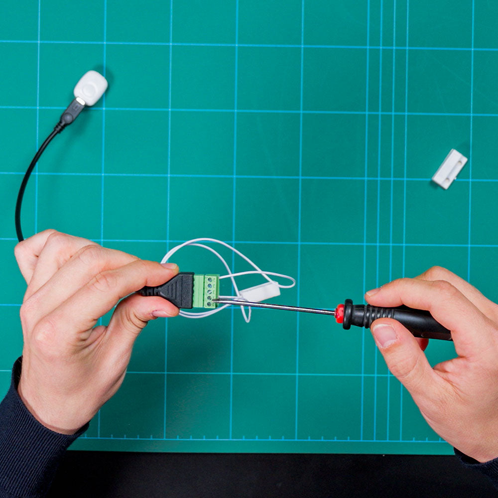 Someone uses a screwdriver to connect wires to the green terminal block of the Shelly USA Micro USB to 5 Terminal Add (Shelly Pill) on a blue cutting mat. A sensor with a white cable and connector is also visible.