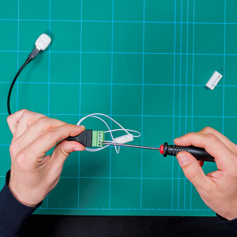 Someone uses a screwdriver to connect wires to the green terminal block of the Shelly USA Micro USB to 5 Terminal Add (Shelly Pill) on a blue cutting mat. A sensor with a white cable and connector is also visible.