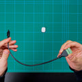 Two hands hold the Shelly USA Micro USB to 5 Terminal Add (Shelly Pill) with green terminal block connector against a green cutting mat background. A small white rectangular object sits on the mat.