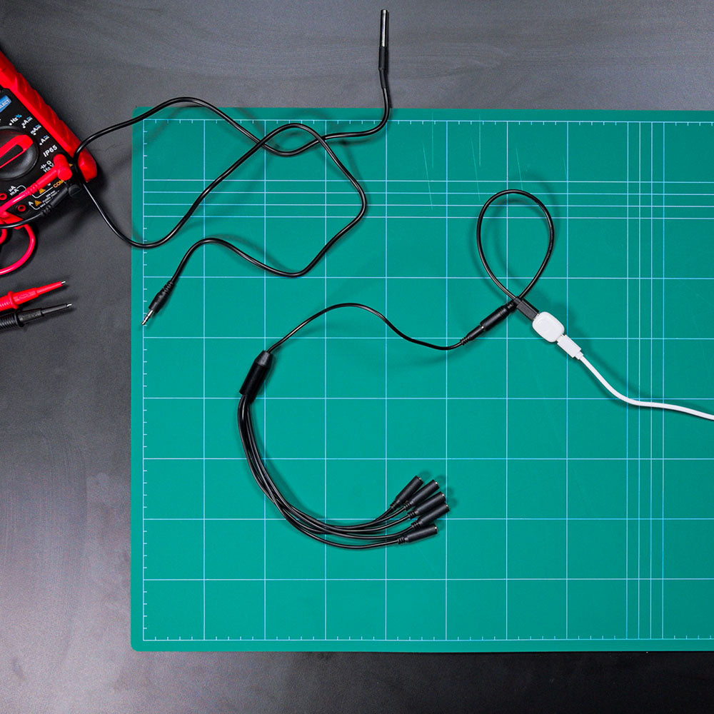 A green cutting mat with grid lines holds a Shelly USA Splitter 1 to 5-3.5 mm Jack (Shelly Pill), surrounded by various electronic cables, alligator clips, and a multimeter with red and black test leads on a dark surface.