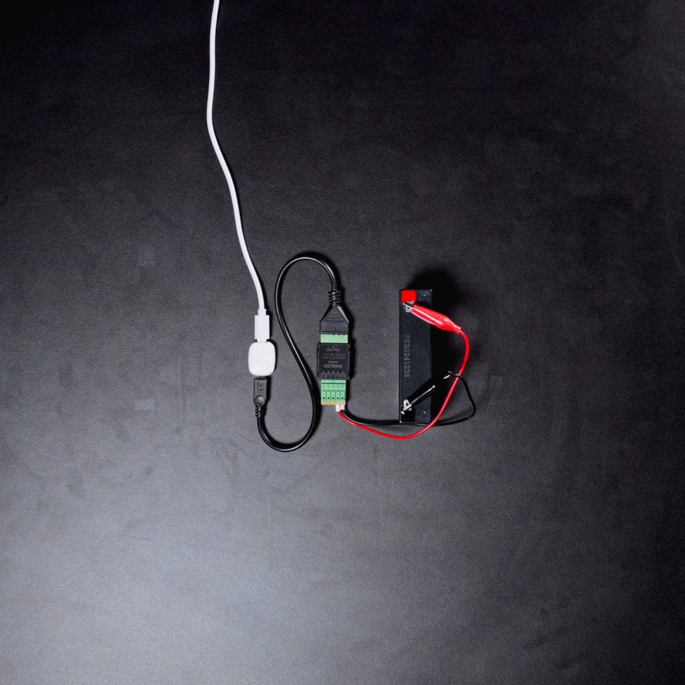 A Shelly USA Analog 0-30V Add-on (Shelly Pill) is connected to a black battery via red and black wires, with a white cable plugged into a USB adapter, all set on a dark surface.