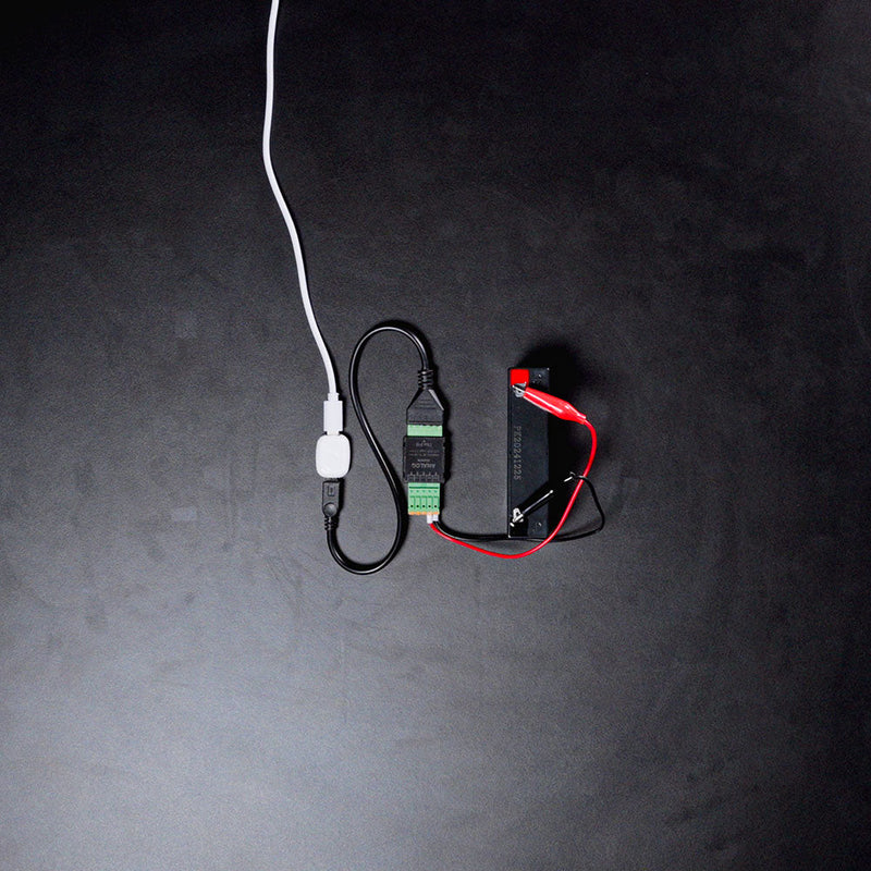 A Shelly USA Analog 0-30V Add-on (Shelly Pill) is connected to a black battery via red and black wires, with a white cable plugged into a USB adapter, all set on a dark surface.