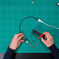 Two hands on a green cutting mat connect wires from a black battery holder to the Shelly USA Analog 0-30V Add-on (Shelly Pill) and a USB cable using alligator clips and connectors.