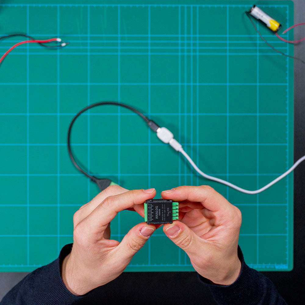 A person holds a Shelly USA Analog 0 - 30V Add-on (Shelly Pill) above a green cutting mat with grid lines, with wires and a USB cable connected to the device.