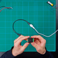 A person holds a Shelly USA Analog 0 - 30V Add-on (Shelly Pill) above a green cutting mat with grid lines, with wires and a USB cable connected to the device.