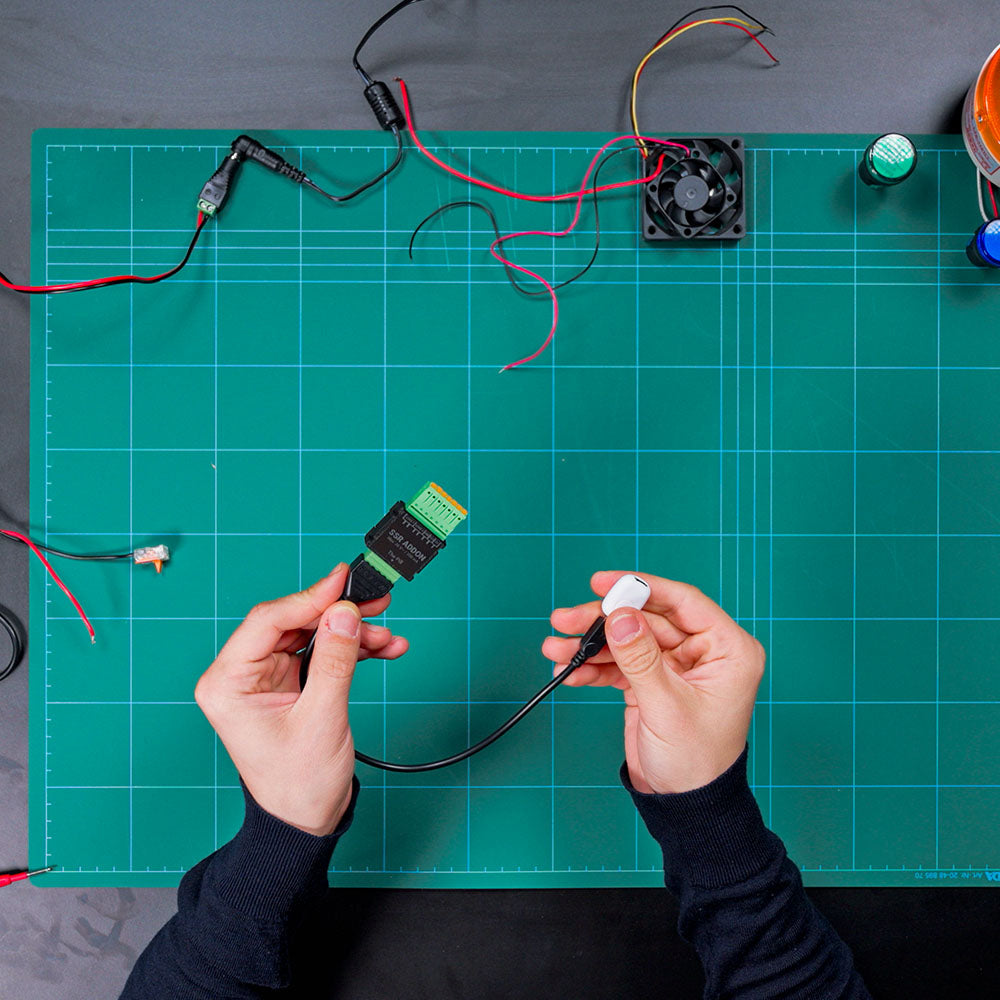 A person holds a Shelly USA SSR Add-on (Shelly Pill) with wires above a green cutting mat, surrounded by loose wires and a small cooling fan on a work table.
