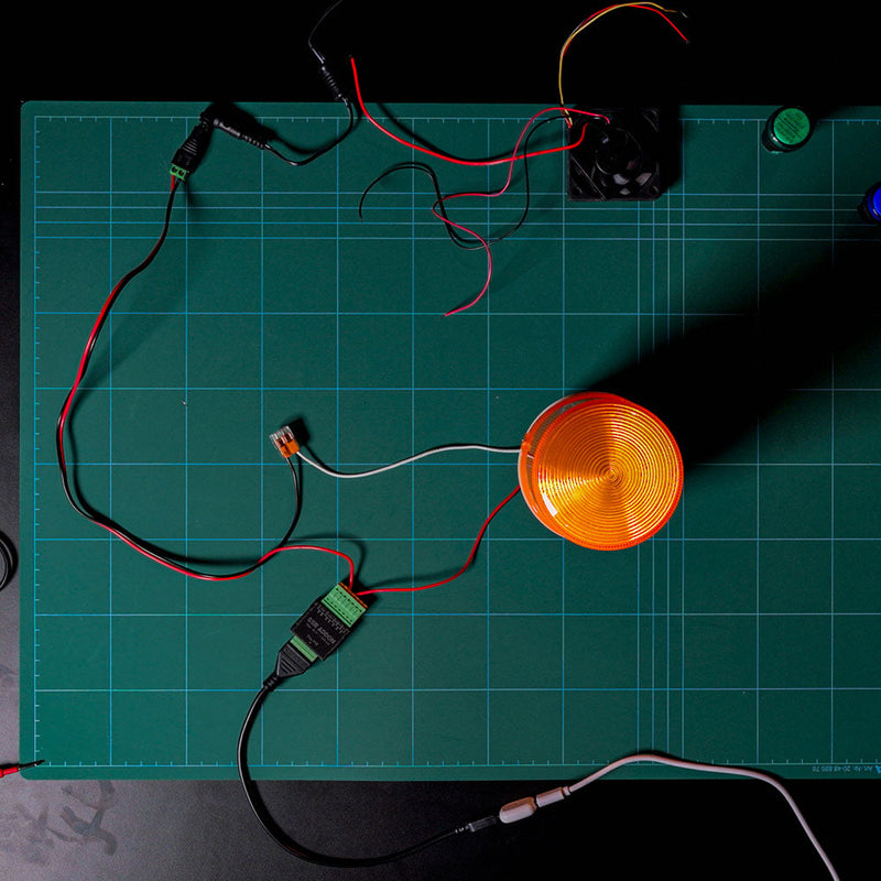 A green cutting mat with grid lines on a table displays the Shelly USA SSR Add-on (Shelly Pill), along with electronic components, wires, a round orange light, and connectors arranged neatly under bright lighting.