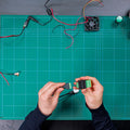 A person’s hands hold the SSR Add-on (Shelly Pill) by Shelly USA above a green cutting mat, surrounded by wires, a fan, and electronics on a workbench.