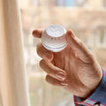 A hand holds the Shelly USA Shelly H&T White, a compact Wi-Fi humidity and temperature sensor with a dial and S symbol on top, near a window with beige curtains.