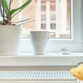 A white mug, a potted plant, and a Shelly USA Shelly H&T White Wi-Fi humidity and temperature sensor rest on a white windowsill in front of a window with blurred buildings outside.