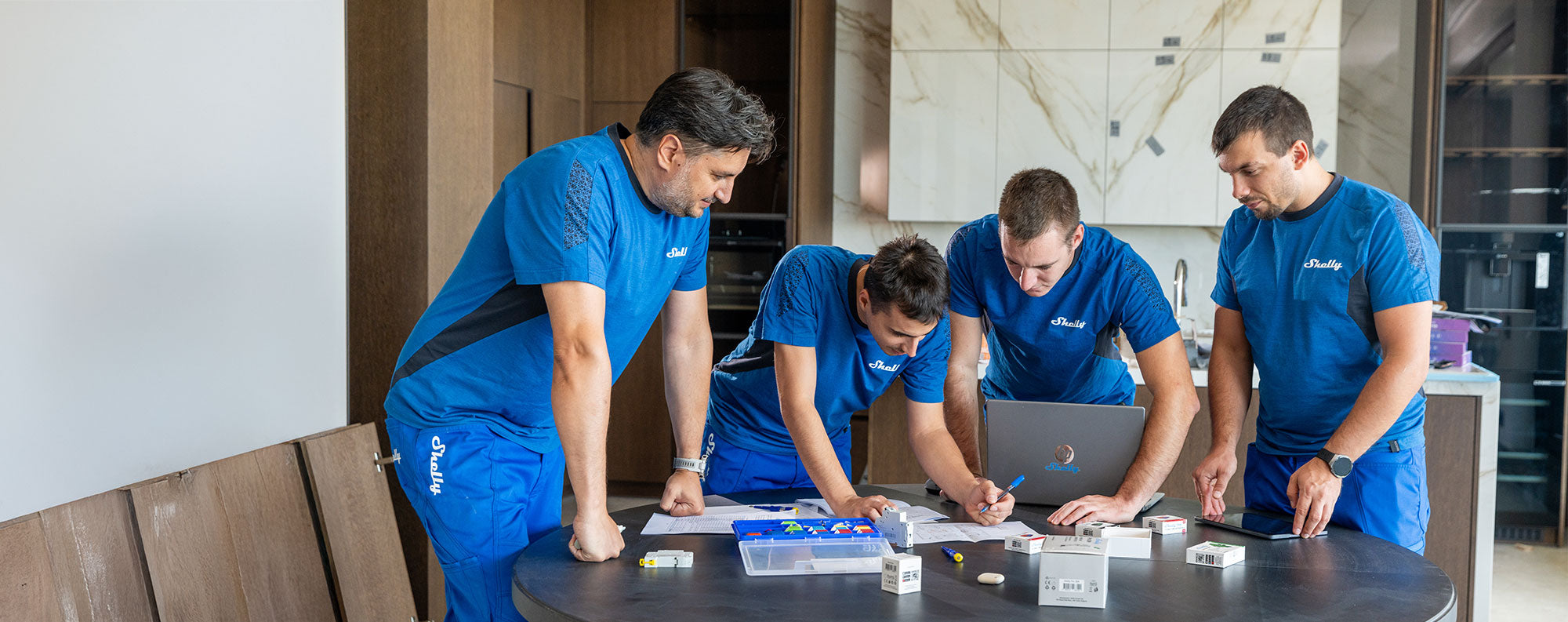 Four men in matching blue uniforms work together at a round table covered with papers, tools, and small electronic devices in a modern, well-lit room with wooden and marble finishes.