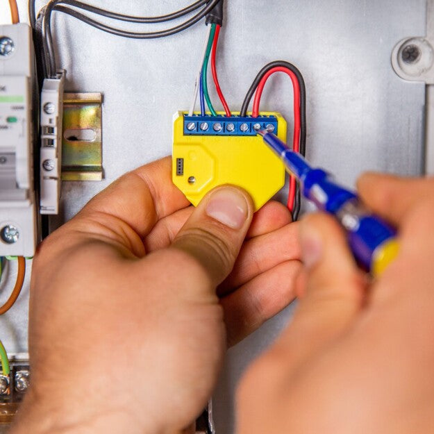 A person uses a screwdriver to connect wires for a Shelly USA Shelly RGBW2 Wi-Fi relay to a yellow electrical terminal block inside an electrical panel.
