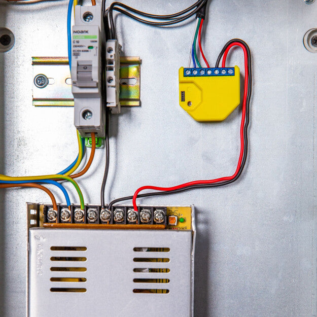 Close-up of a Shelly USA Shelly RGBW2 smart relay module for Wi-Fi and smart home integration, connected with wires alongside a circuit breaker and terminal block inside a metal electrical enclosure.
