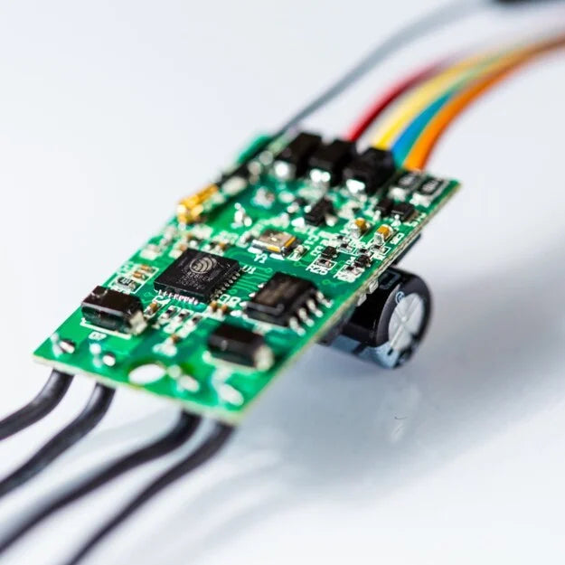 Close-up of a Shelly Uni by Shelly USA, an electronic circuit board with various components, colorful wires, a capacitor, and a smart Wi-Fi module for sensor control, set against a white background.