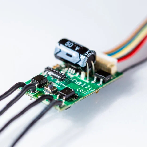 Close-up of the Shelly Uni by Shelly USA, a compact electronic circuit board featuring black and silver components, a cylindrical capacitor, and colored wires, engineered for data measurement in smart Wi-Fi modules.