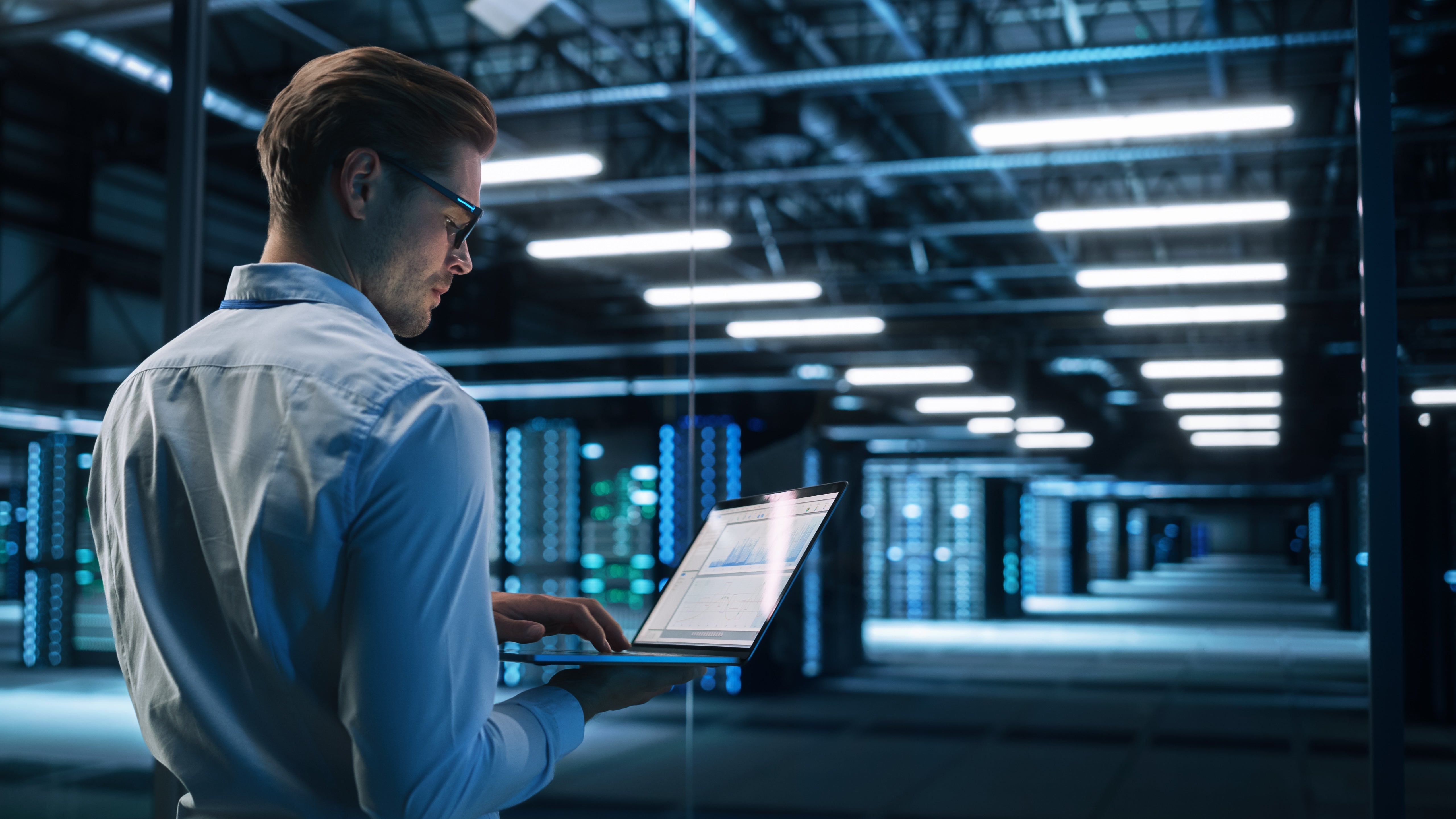 A man in glasses and a white shirt uses a laptop while standing in a modern server room filled with rows of illuminated server racks and overhead lights.