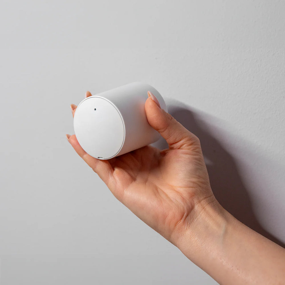 A hand with manicured nails holds the Shelly BLU TRV by Shelly USA, a compact cylindrical WiFi radiator valve with a smooth surface and a small hole near the edge, against a light gray wall.